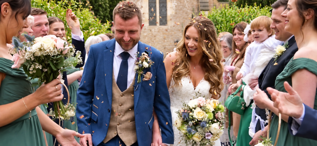 Jess and Callum smiling beneath confetti with their wedding guests outside St Mary’s Church at Orchardleigh