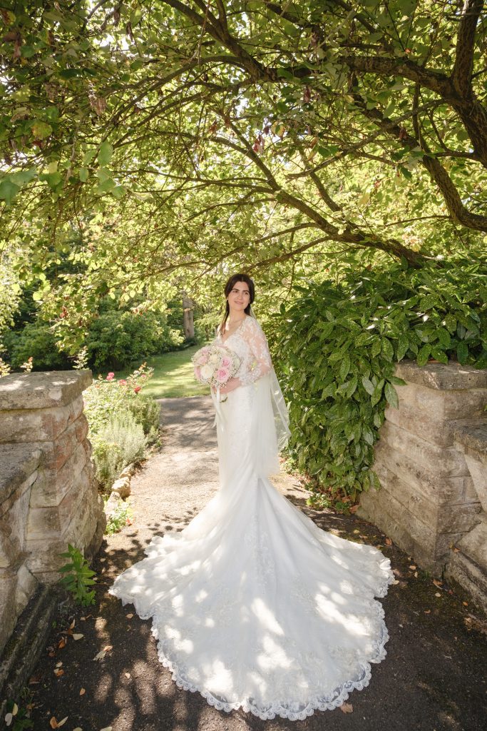 Elaina standing beneath leafy branches beside a stone wall, with sunlight falling across the lace train of her wedding dress.
