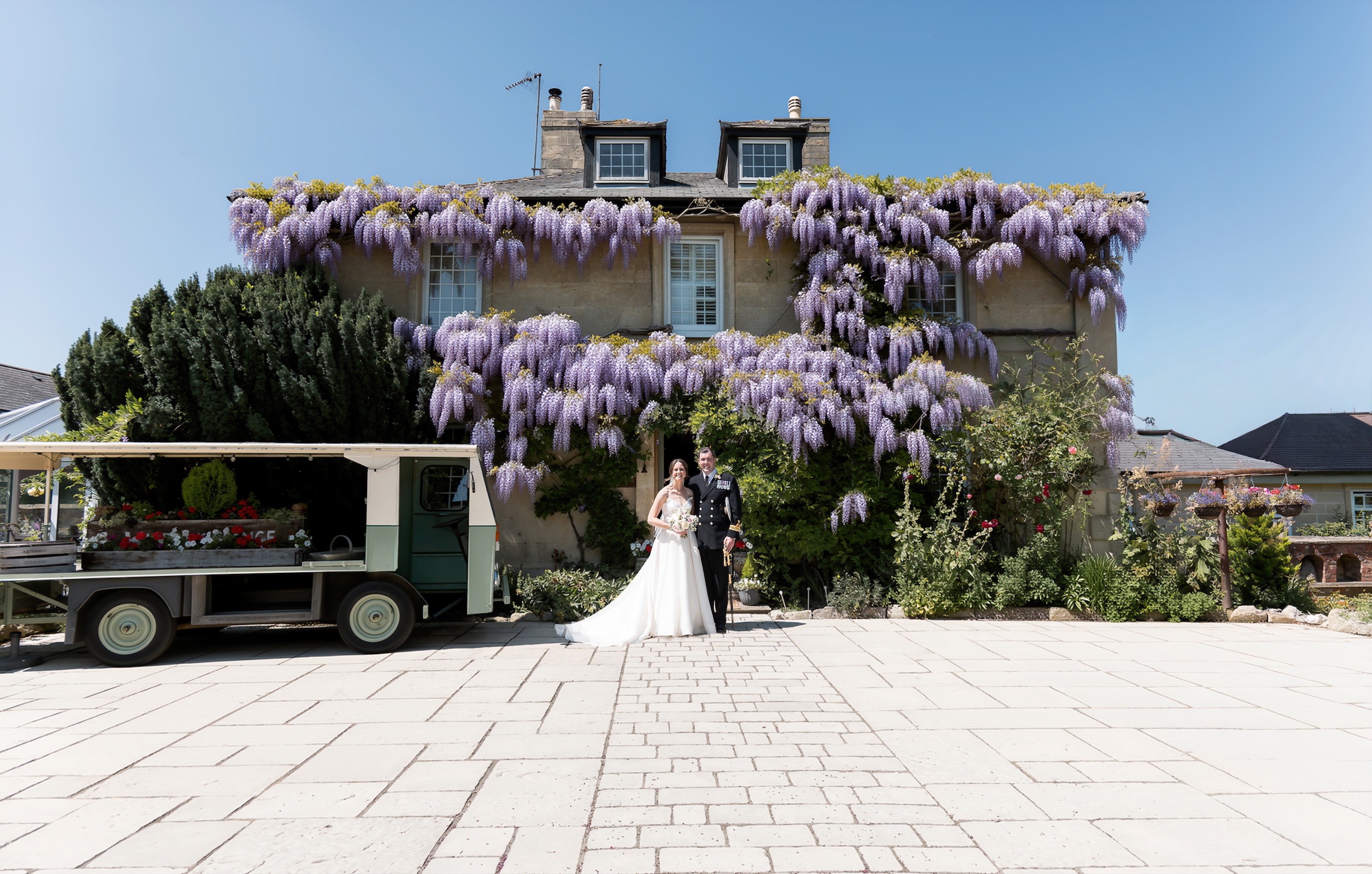 Laura and Ross beneath flowering wisteria outside Widbrook Grange, beside a vintage vehicle filled with flowers.
