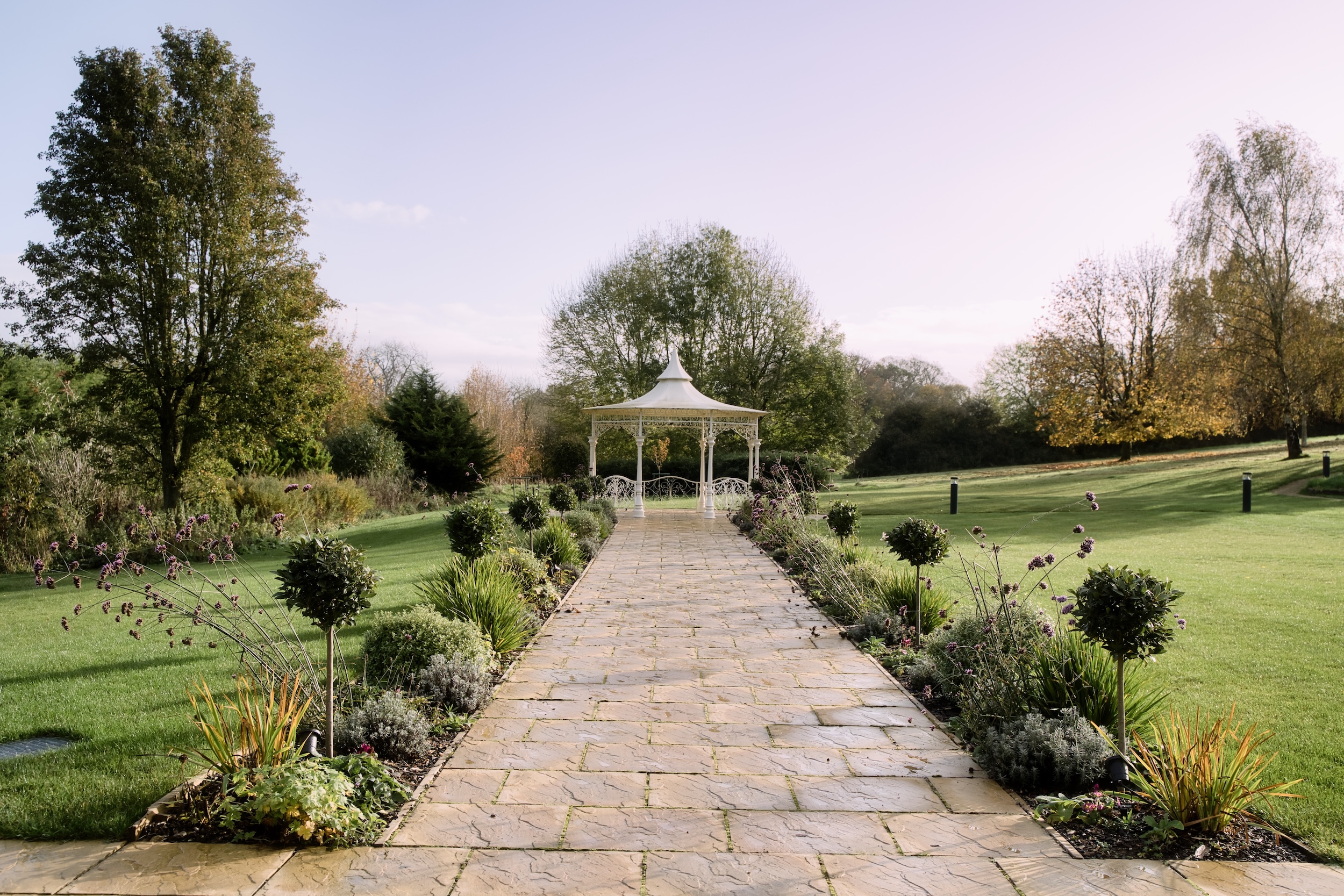 A stone path bordered by flowers and clipped shrubs leading to the ornate white pavilion in The Pear Tree gardens.