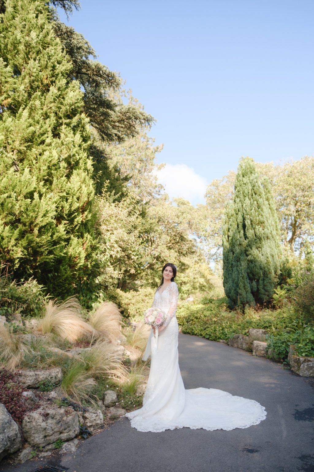 Elaina holding her pink and ivory bouquet on a path in Bath’s Botanical Gardens, with her lace train spread behind her.