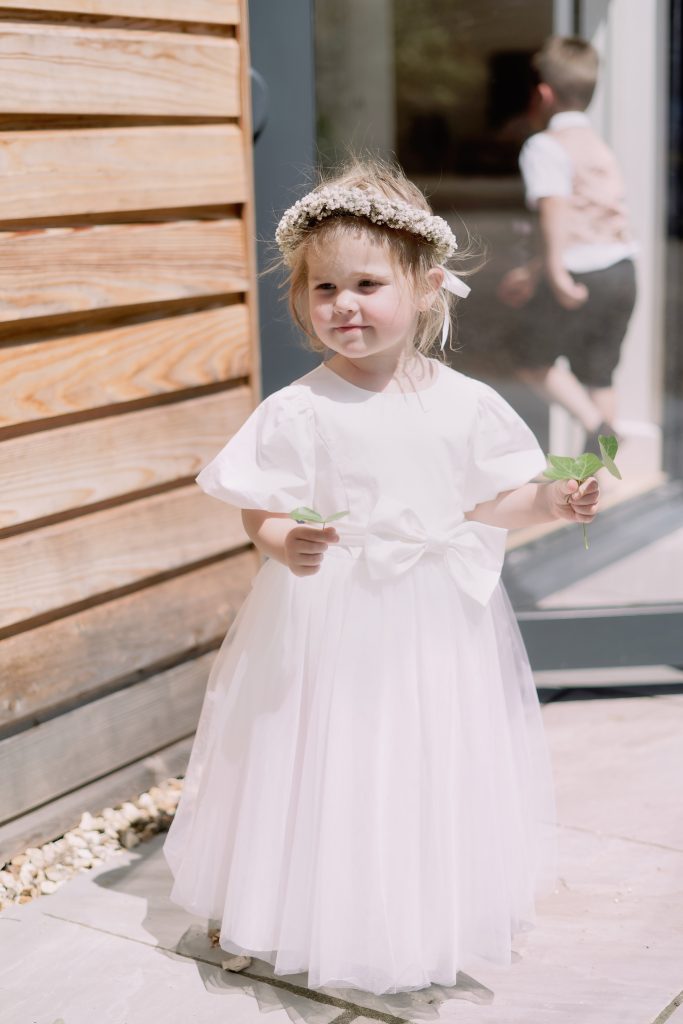 A young flower girl wearing a pale pink dress and a flower crown, holding leaves beside a wooden wall.