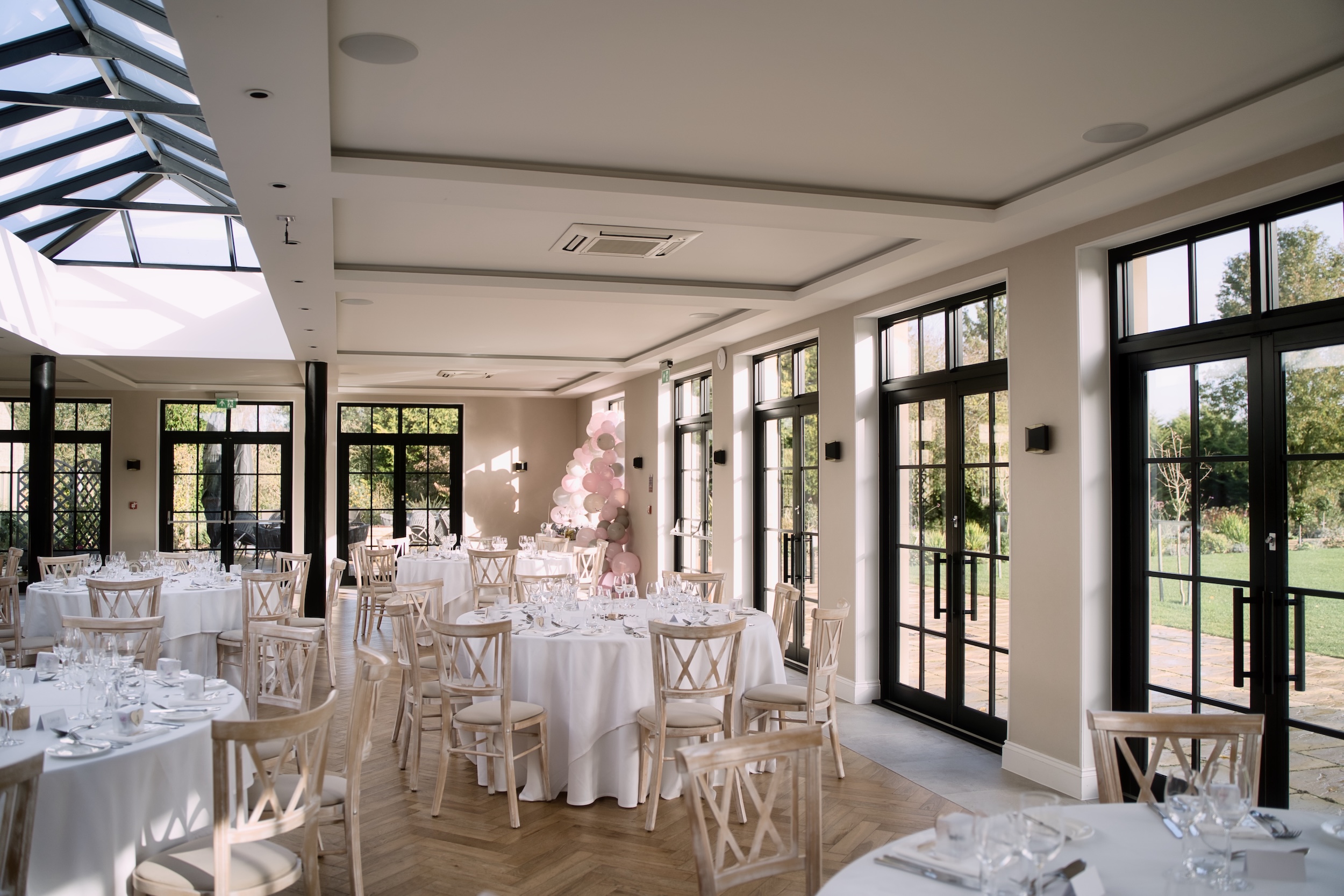 White-clothed wedding tables and pale pink decorations beneath the glass roof and tall windows of The Pear Tree’s Orangery.