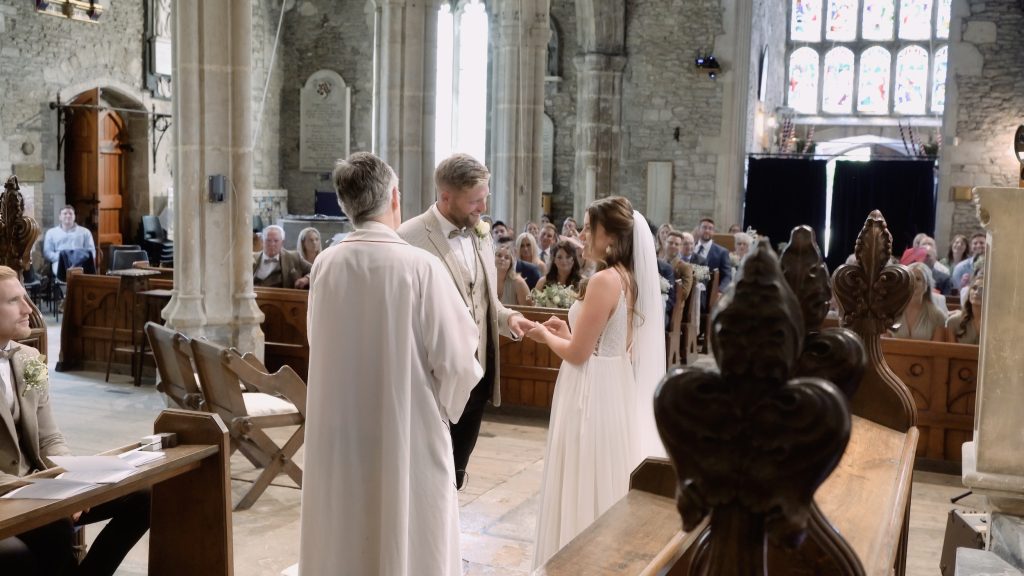 Alice and Ben holding hands during their wedding ceremony at St George’s Church in Beckington, with the vicar and guests nearby.
