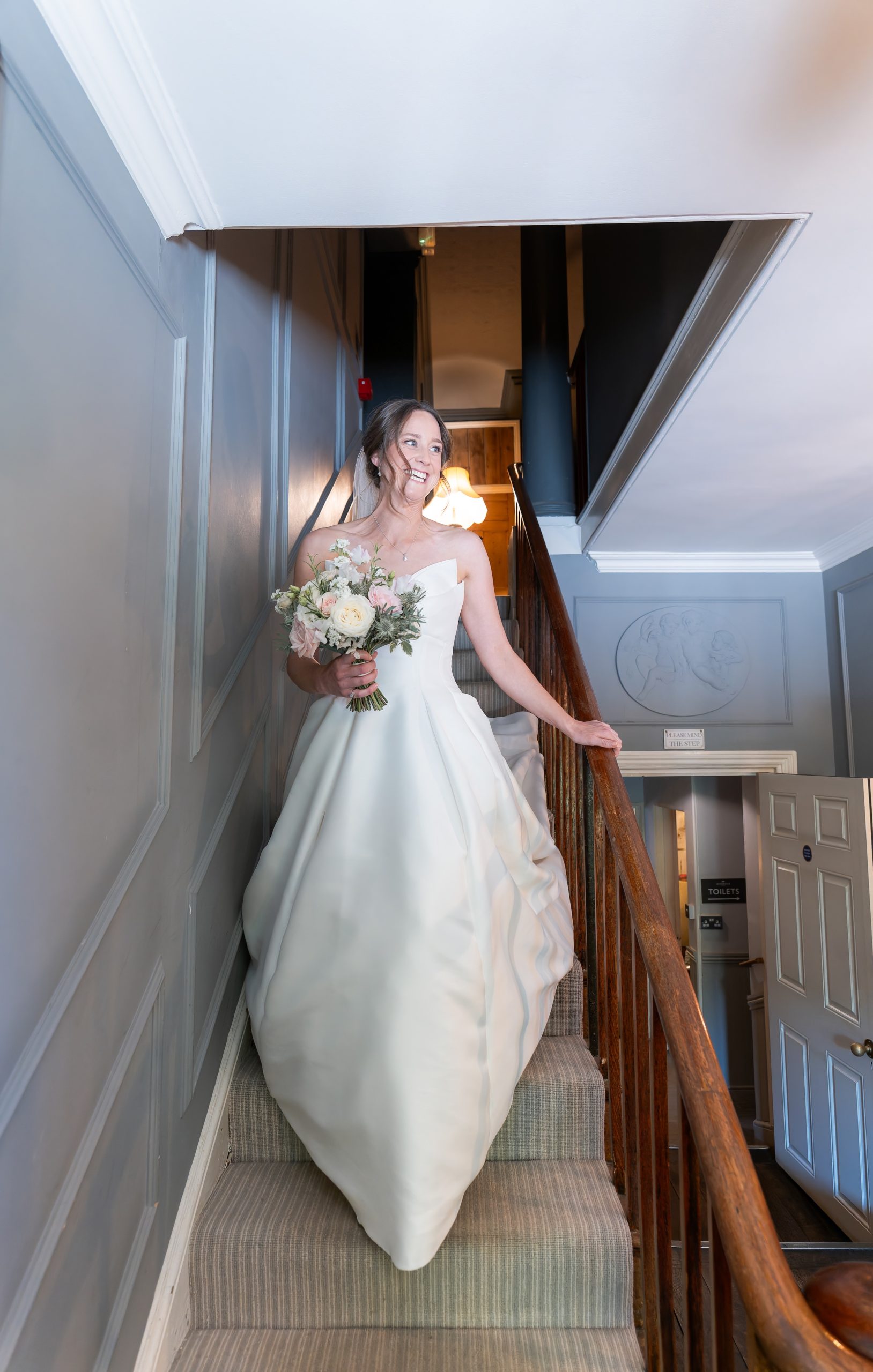 Laura in her wedding dress holding a pale flower bouquet on a wooden staircase at Widbrook Grange.