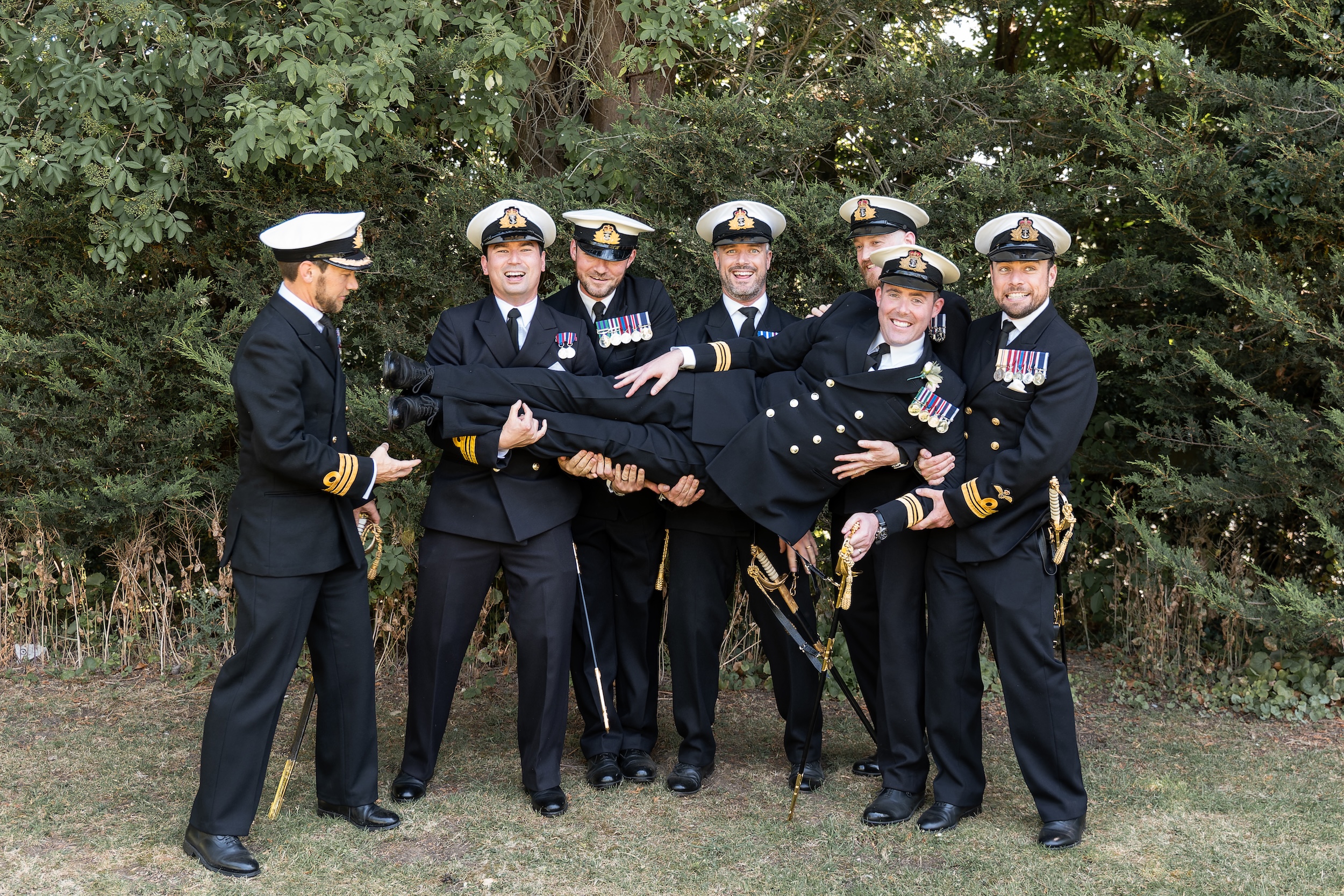 Members of Laura and Ross’s wedding party in military dress uniform lifting a friend and laughing in the gardens at Widbrook Grange.