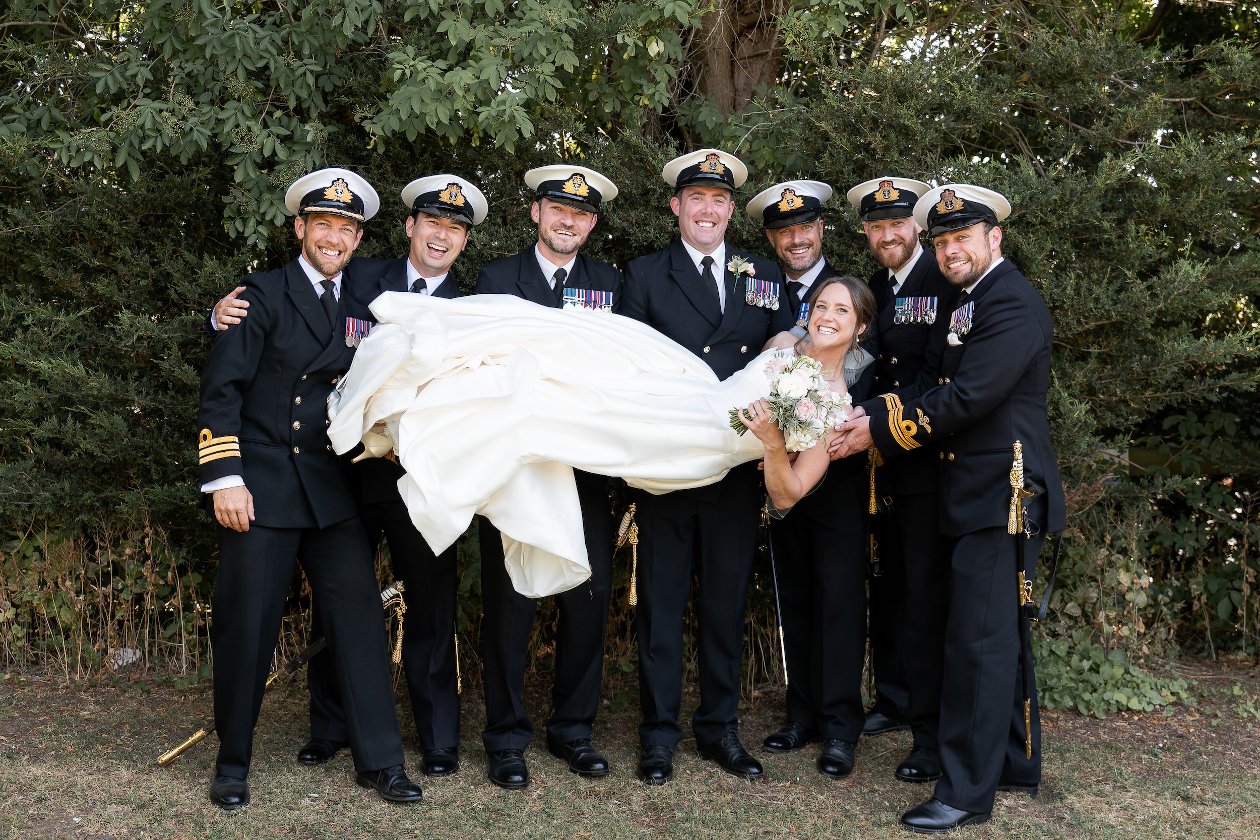 Laura laughing as Ross and the wedding party lift her for a playful group photograph in the gardens at Widbrook Grange.