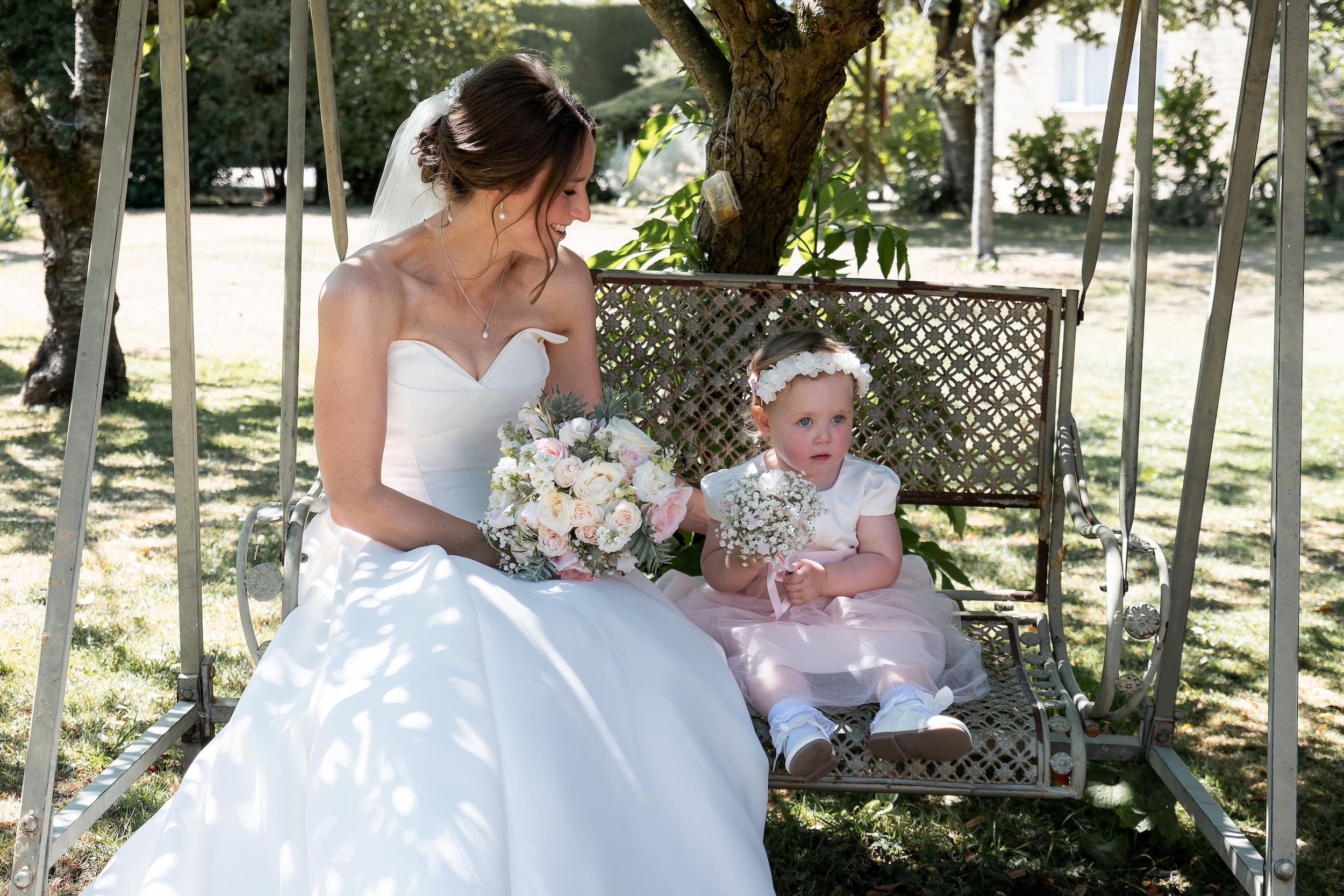 Laura sitting with a young flower girl on a metal garden swing beneath a tree at Widbrook Grange.
